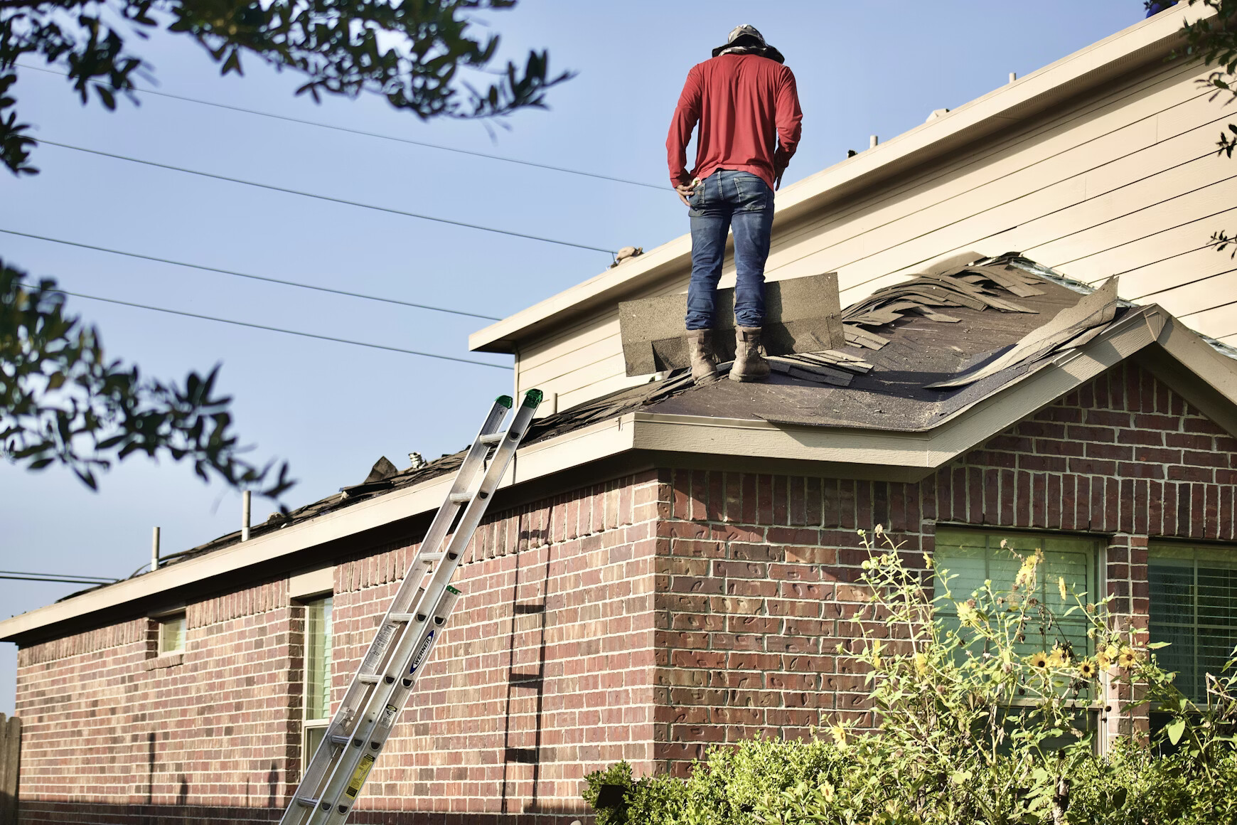 A construction worker wearing a yellow hard hat, safety goggles, and an orange high-visibility vest uses a power drill to secure grey roof tiles on a bright, sunny day.