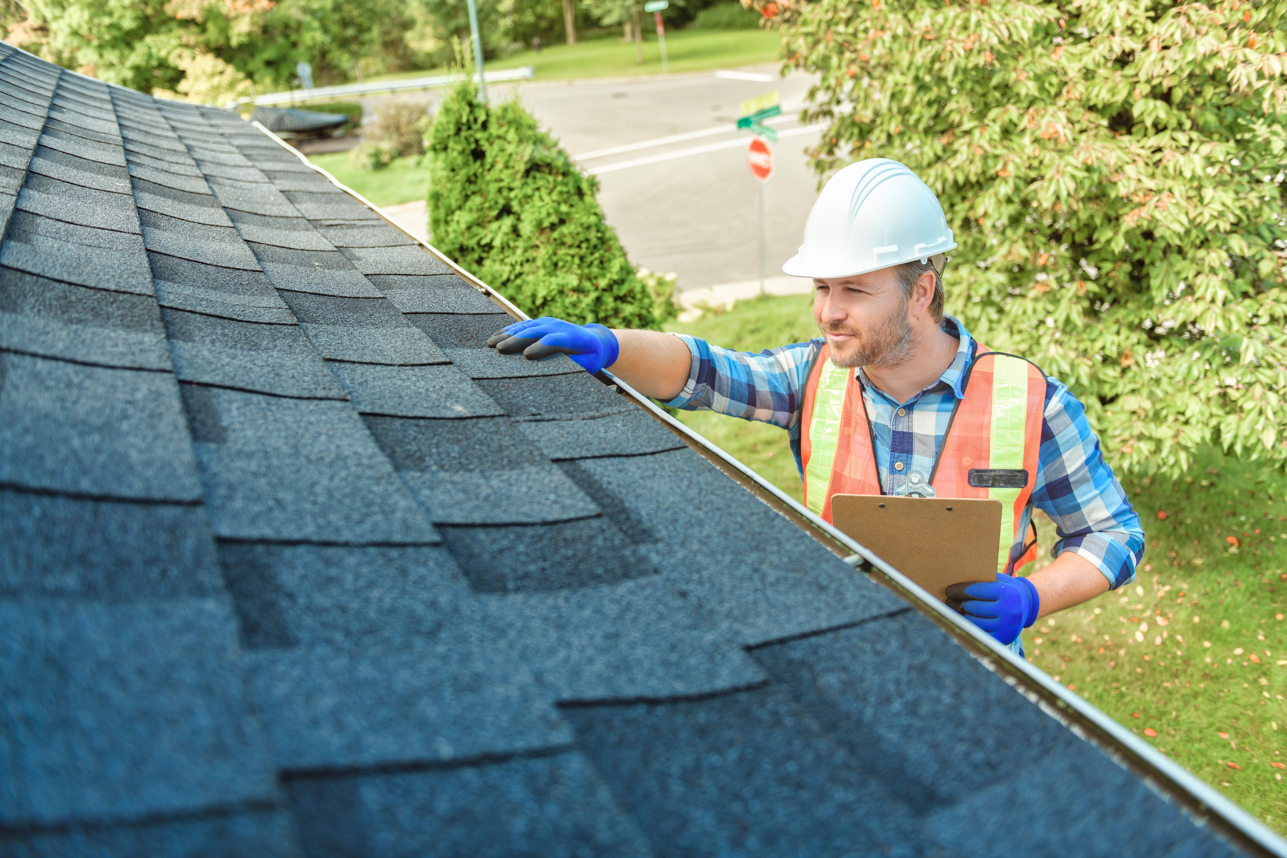 Roofing professional wearing a hard hat and safety vest inspecting asphalt shingles on a residential roof while holding a clipboard during an exterior roof assessment.