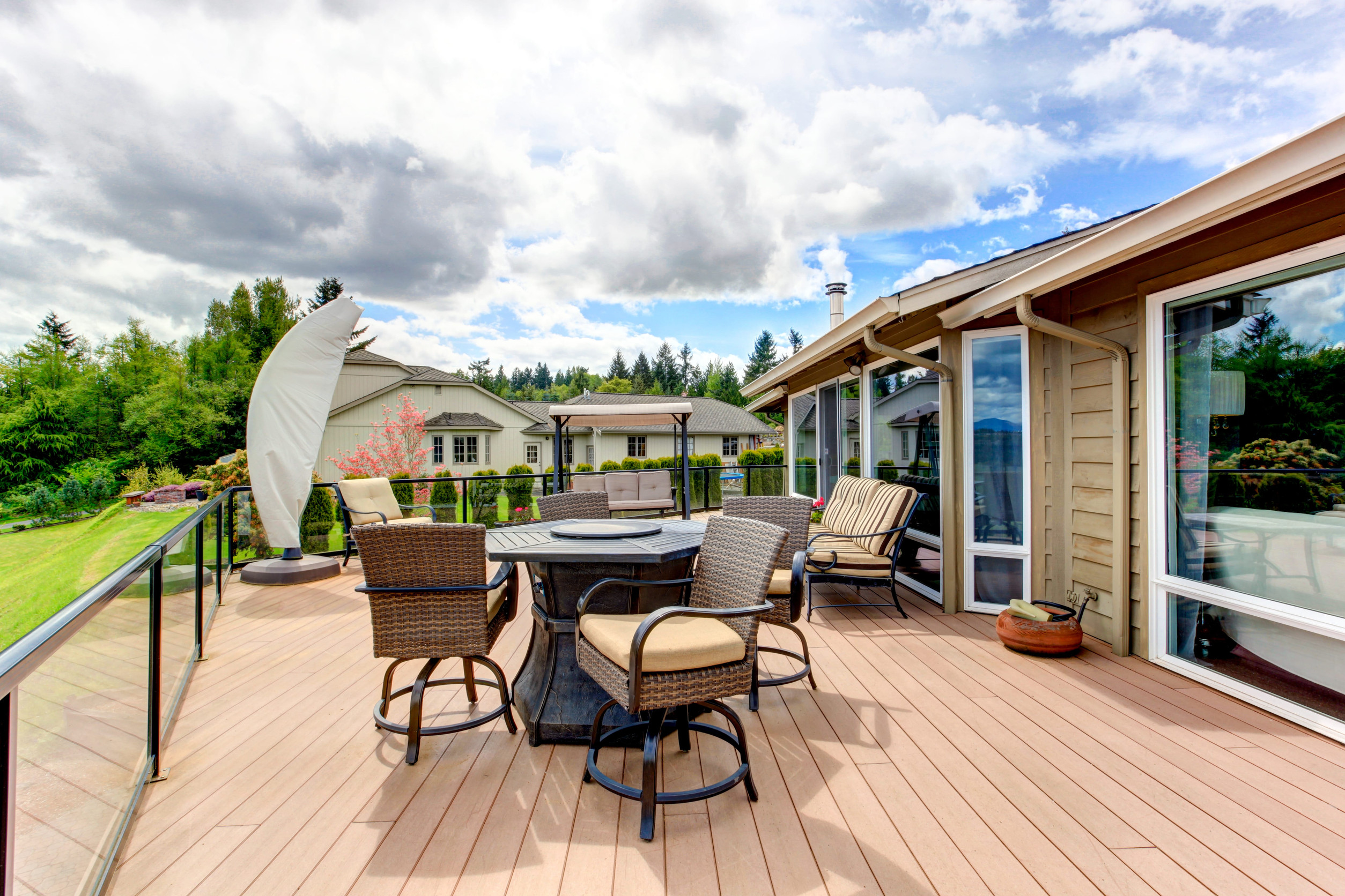 30130228_m A spacious, well-maintained backyard deck attached to a home features outdoor dining furniture, cushioned seating, glass railings, and scenic landscaping, creating a safe and inviting space for outdoor living and entertaining.
